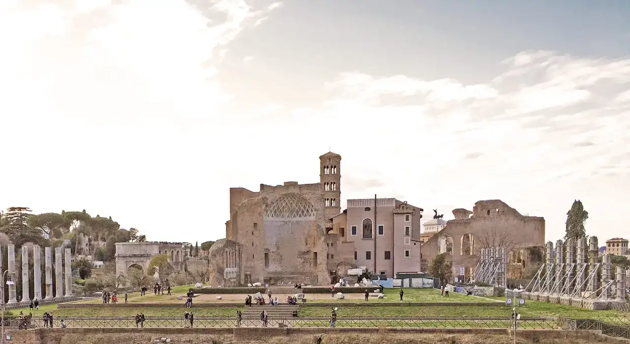 Resti del Foro Romano, con il **tempio di Venere Roma** in lontananza, sotto un cielo nuvoloso. Turisti passeggiano tra le rovine.