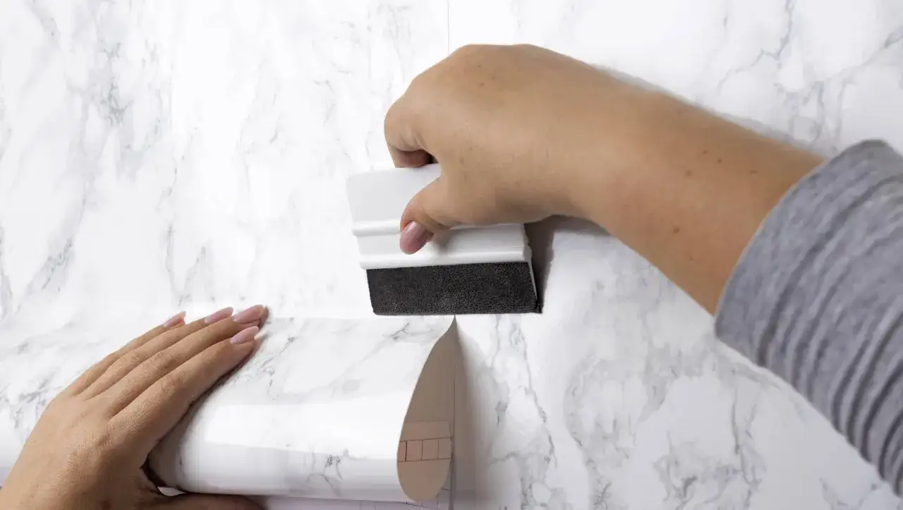 Manos aplicando papel tapiz con dise&ntilde;o de m&aacute;rmol a una pared. Herramienta para alisar burbujas. Gu&iacute;a visual de c&oacute;mo empapelar una pared.