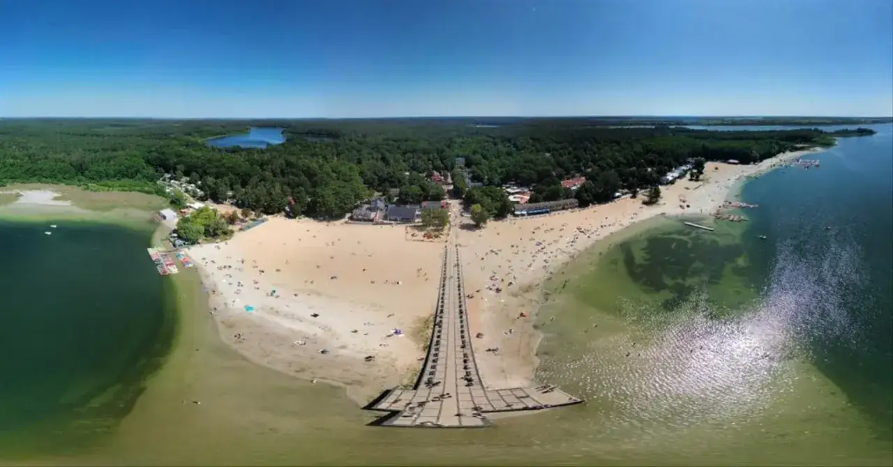 Słoneczny dzień nad Skorzęcinem. Tłumy plażowiczów, molo i las otaczają turkusowe jezioro.