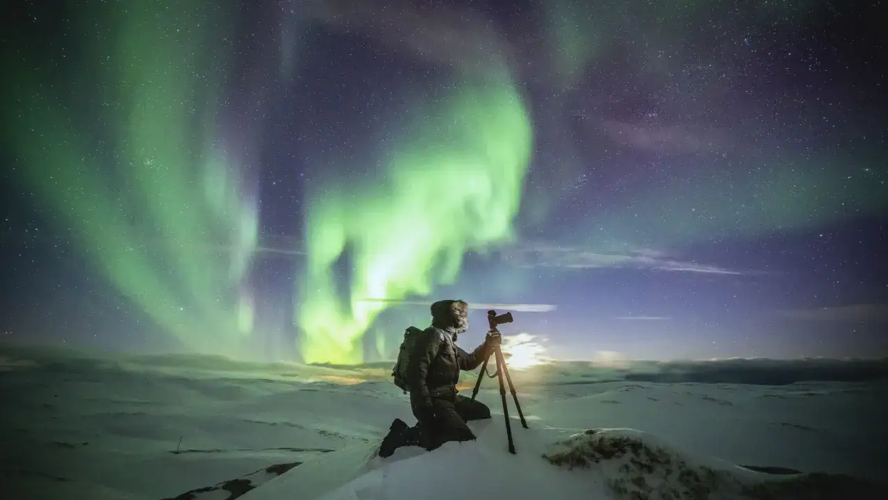 Un photographe, armé de son trépied, capture la magie des aurores boréales vertes et violettes dans un paysage enneigé.
