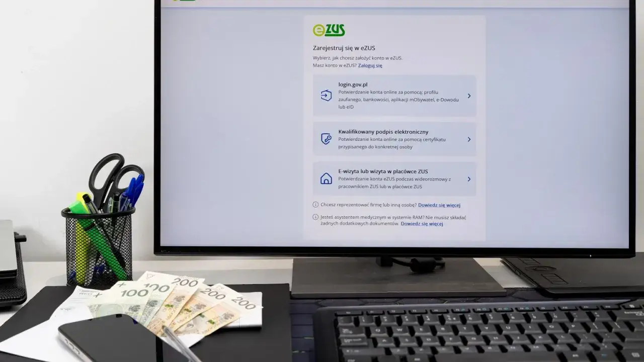 Na biurku leżą pieniądze, telefon i artykuły biurowe. Na monitorze widać stronę logowania do systemu eZUS, gdzie można załatwić sprawy związane z zus 500 plus.