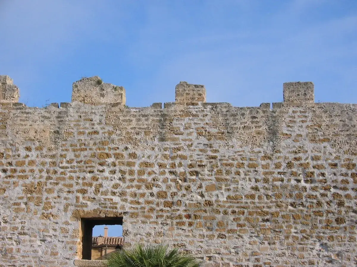 En gammal stenmur med krenelering och ett litet fönster, som en del av den historiska marknad Alcudia.