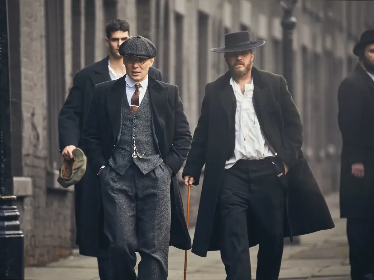 Uomini in abiti eleganti, con cappelli e cappotti, camminano con passo deciso, incarnando lo stile Peaky Blinders.