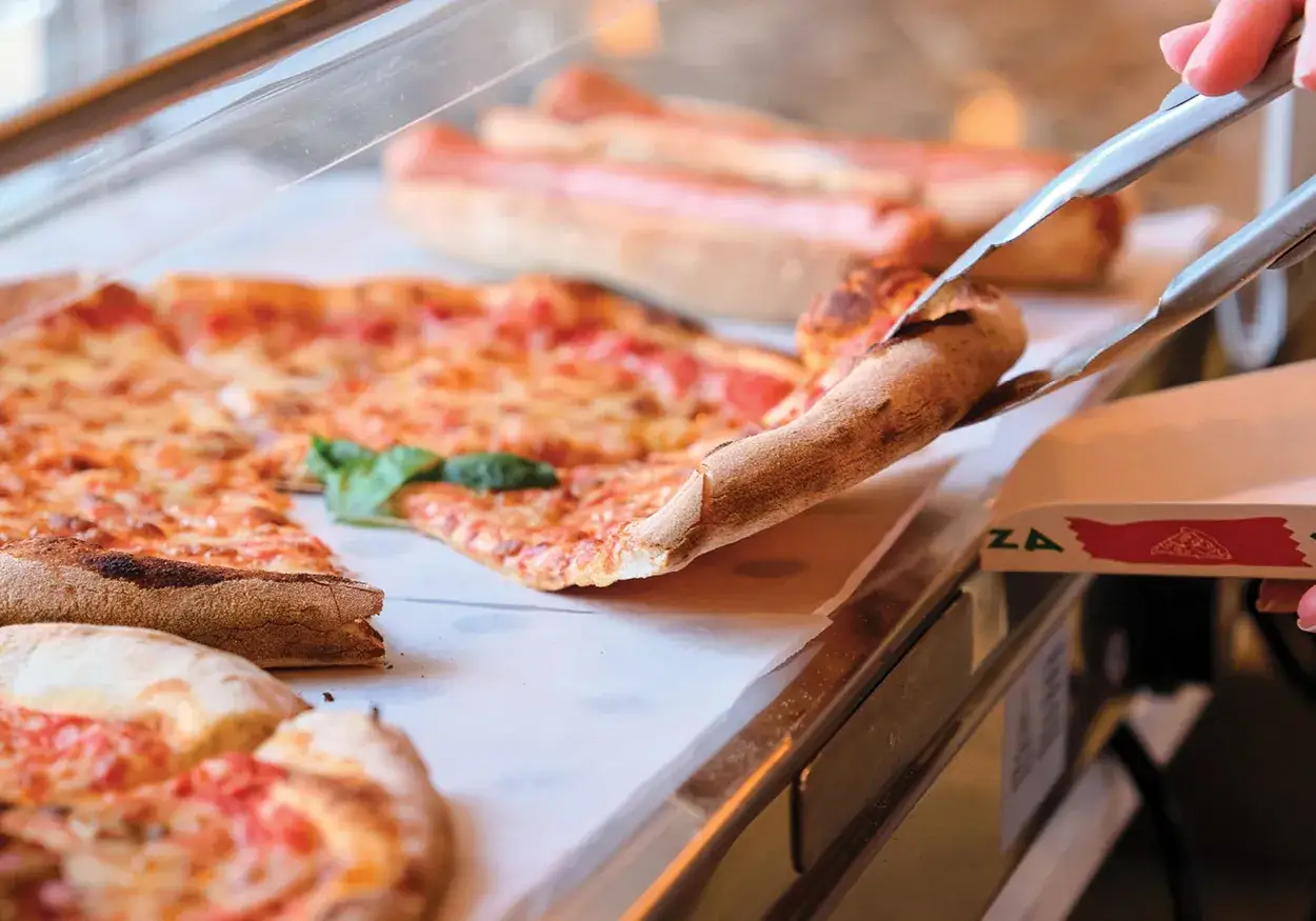 Eine Zange hebt ein St&uuml;ck New York Style Pizza aus der Auslage. Der K&auml;se schmilzt, die Kruste ist goldbraun.