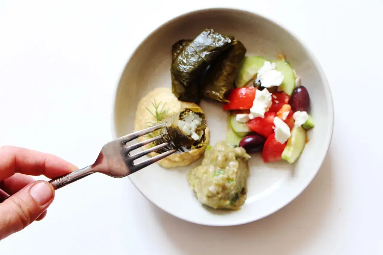 Une fourchette soul&egrave;ve une feuille de vigne farcie, &agrave; c&ocirc;t&eacute; d'une salade grecque et de houmous.
