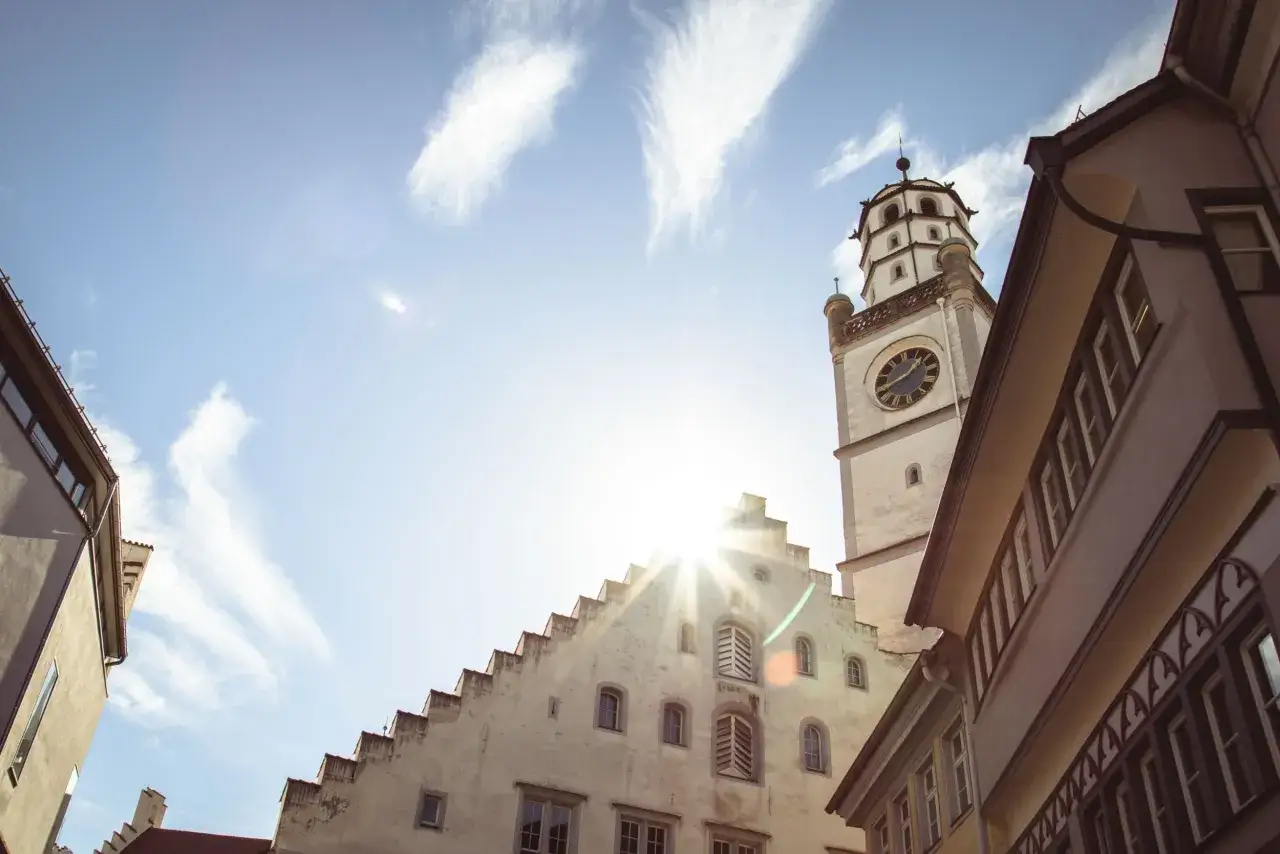 Der Blaserturm Ravensburg ragt unter blauem Himmel mit Federwolken empor. Sonnenschein bricht durch die Architektur.