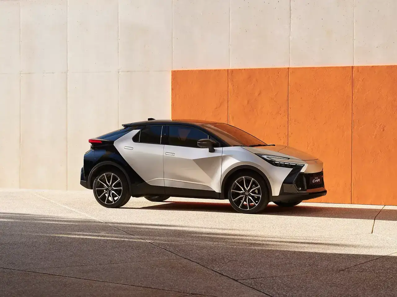 En stilren Toyota C-HR, ett exempel p&aring; bra hybridbilar, parkerad framf&ouml;r en modern v&auml;gg i orange och beige.