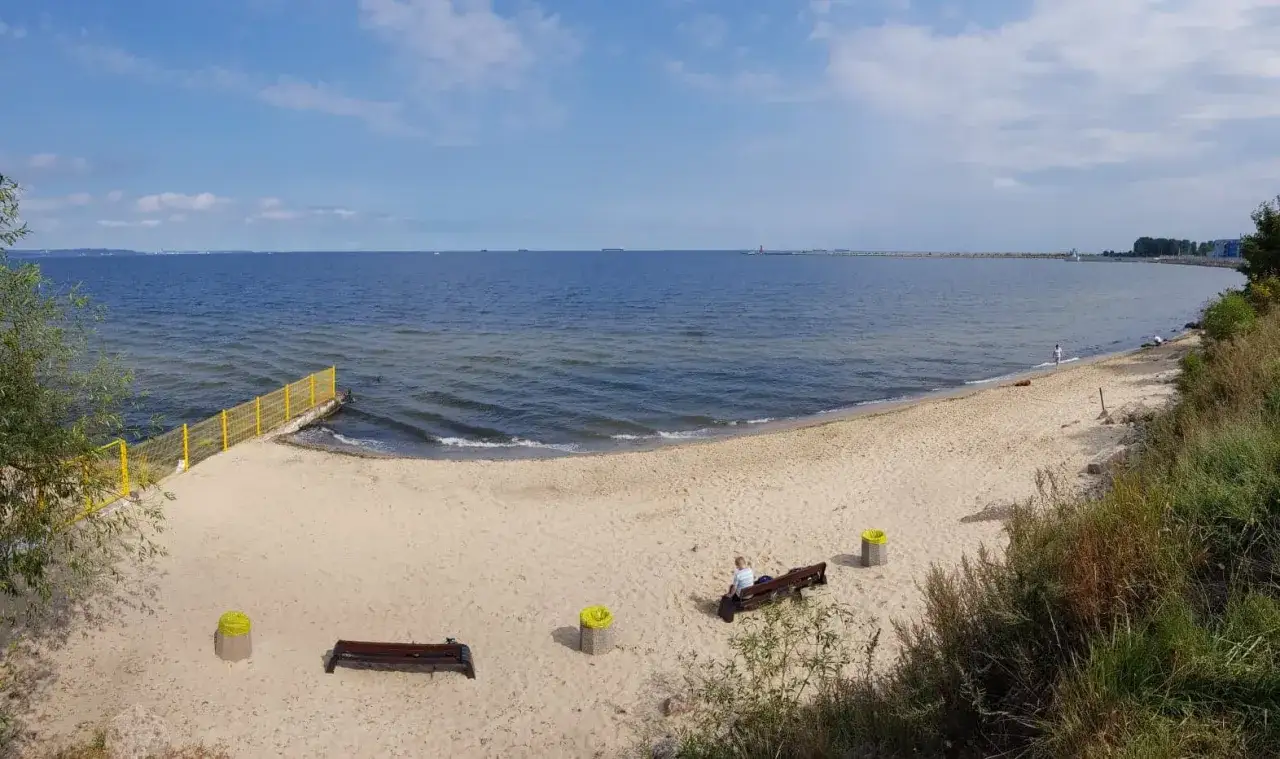 Plaża dla psów z łagodnym zejściem do morza. Na piasku ławki i kosze. W oddali widać falochron i statki.