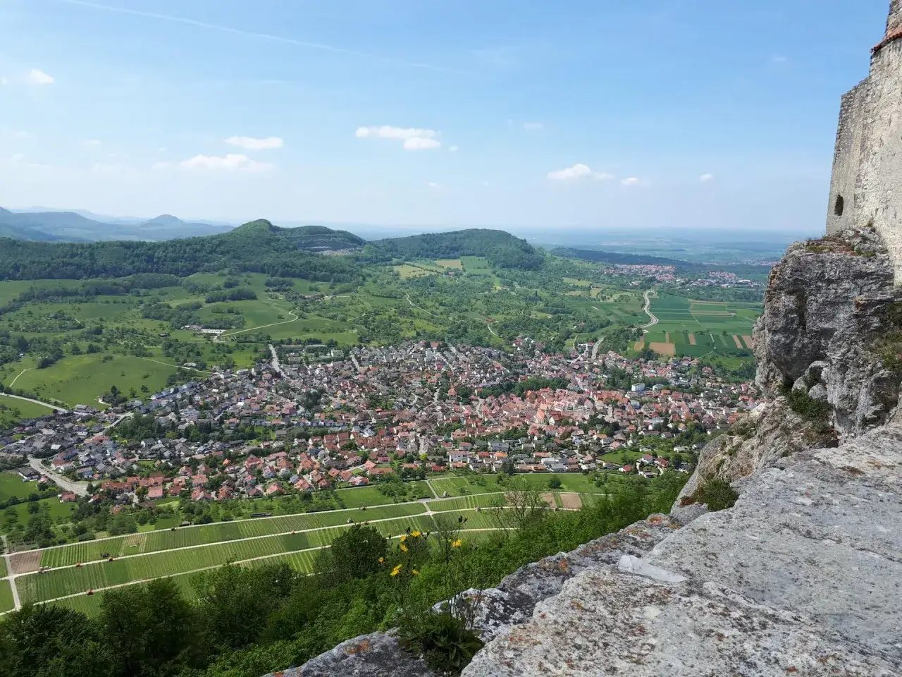 Blick von einer Felswand auf die **Ruine Hofen** und das darunterliegende Dorf mit Weinbergen und gr&uuml;nen H&uuml;geln.