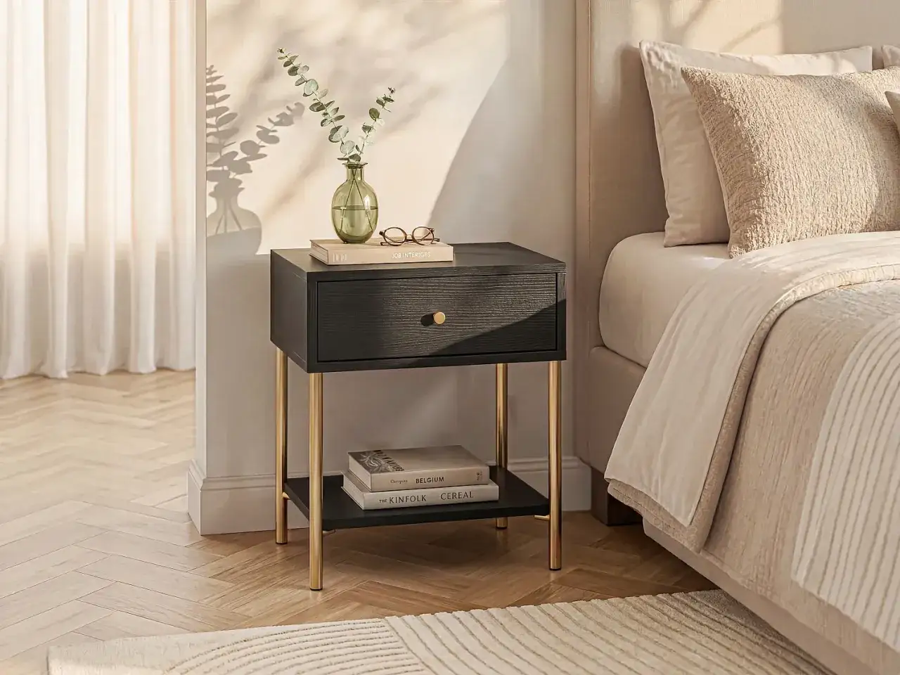 Ett elegant svart sängbord med guldfötter står bredvid en inbjudande säng. En grön vas med eukalyptus och böcker pryder bordet, vilket skapar en lugn atmosfär i det annars svart sovrummet.