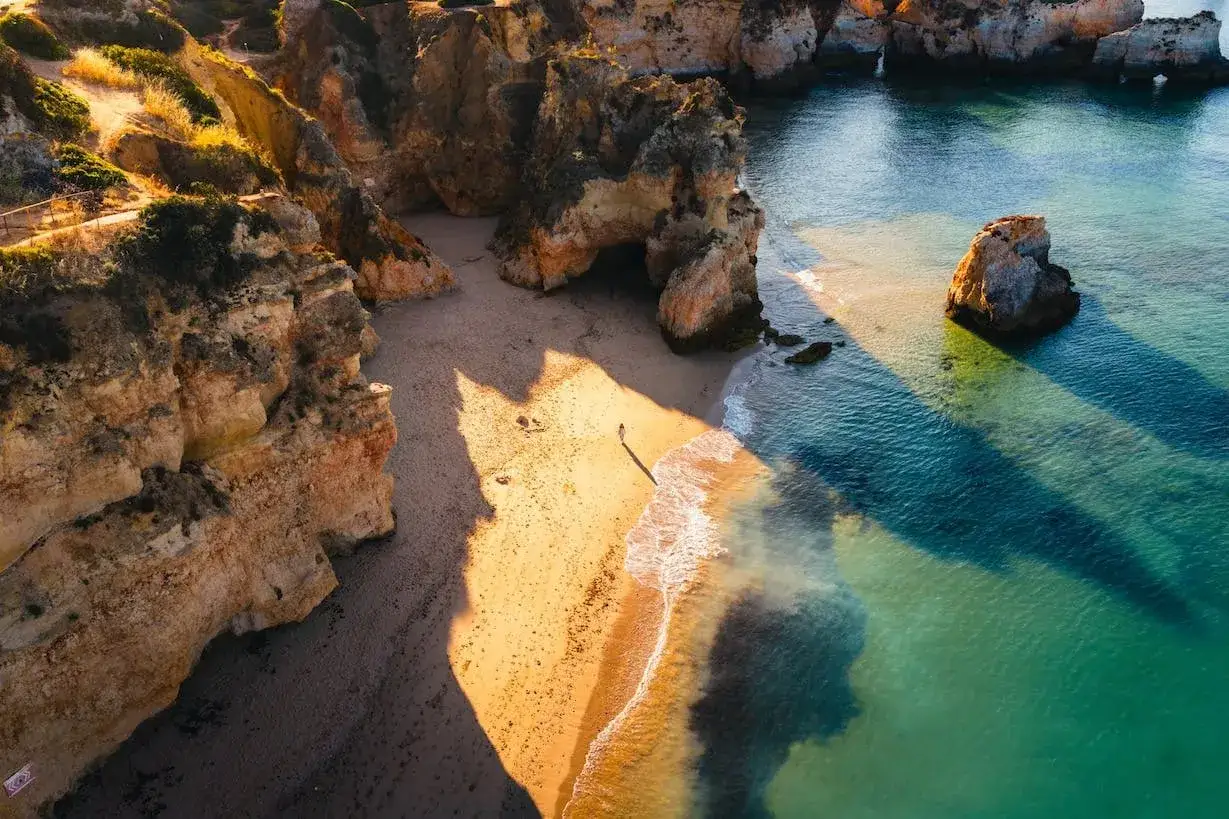 Una persona camina por una playa dorada en las **playas Algarve**, con acantilados imponentes y aguas turquesas.