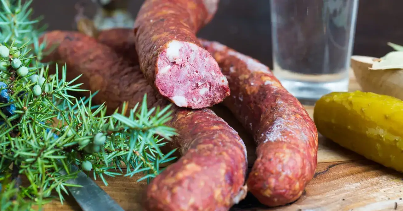 Kiełbasa z białym nalotem, gałązka jałowca, ogórek kiszony i szklanka wódki na drewnianej desce.