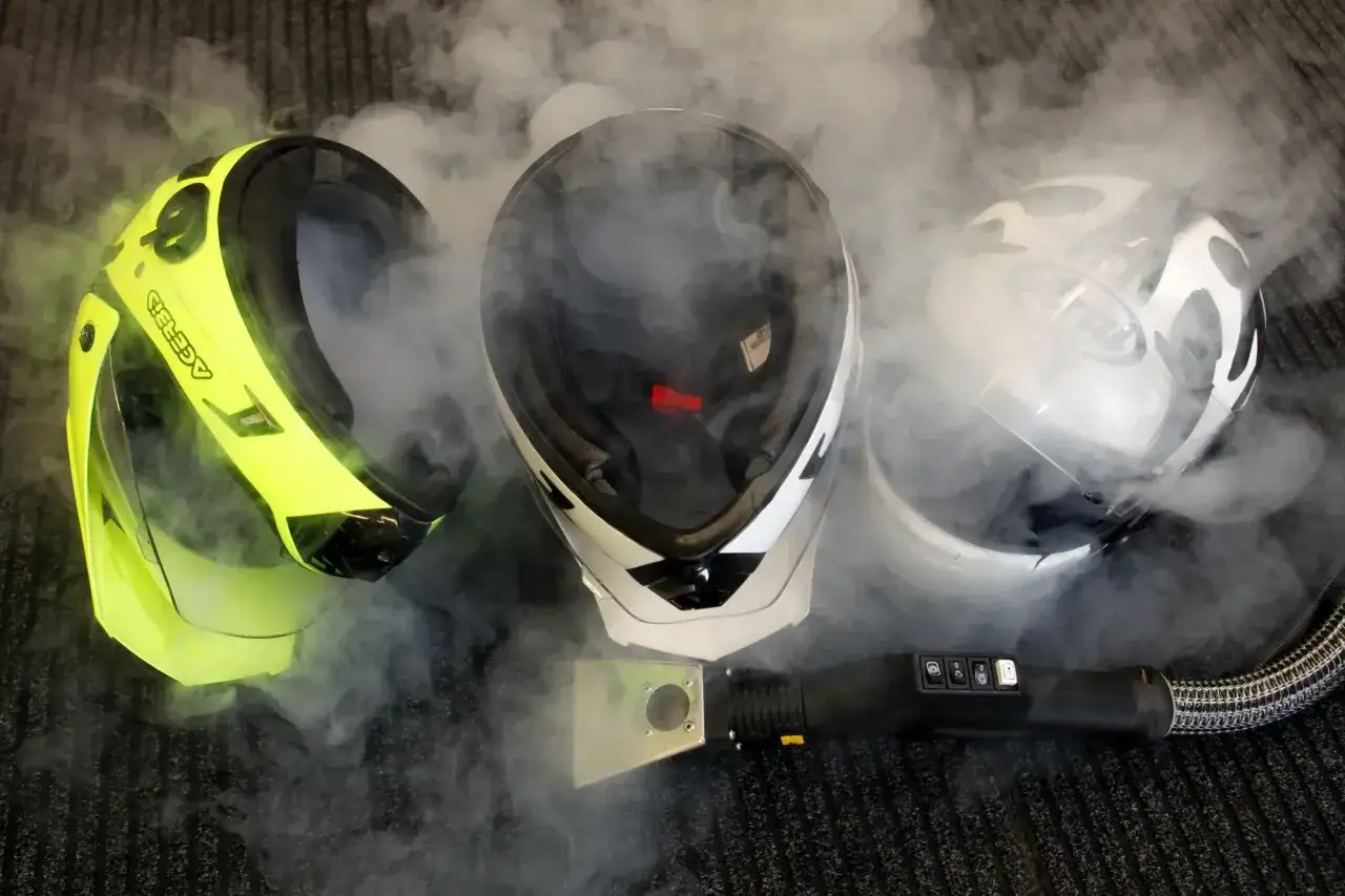 Tre caschi, uno giallo fluo, uno bianco e nero, uno bianco, circondati da vapore. Uno strumento di pulizia a vapore &egrave; posizionato davanti. Ricorda, il casco non deve essere pulito con solventi.