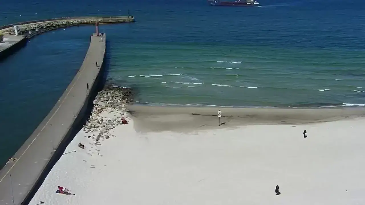 Spacer spaceruje po molo w Łebie, podczas gdy na plaży odpoczywają ludzie. Na morzu widać statek.