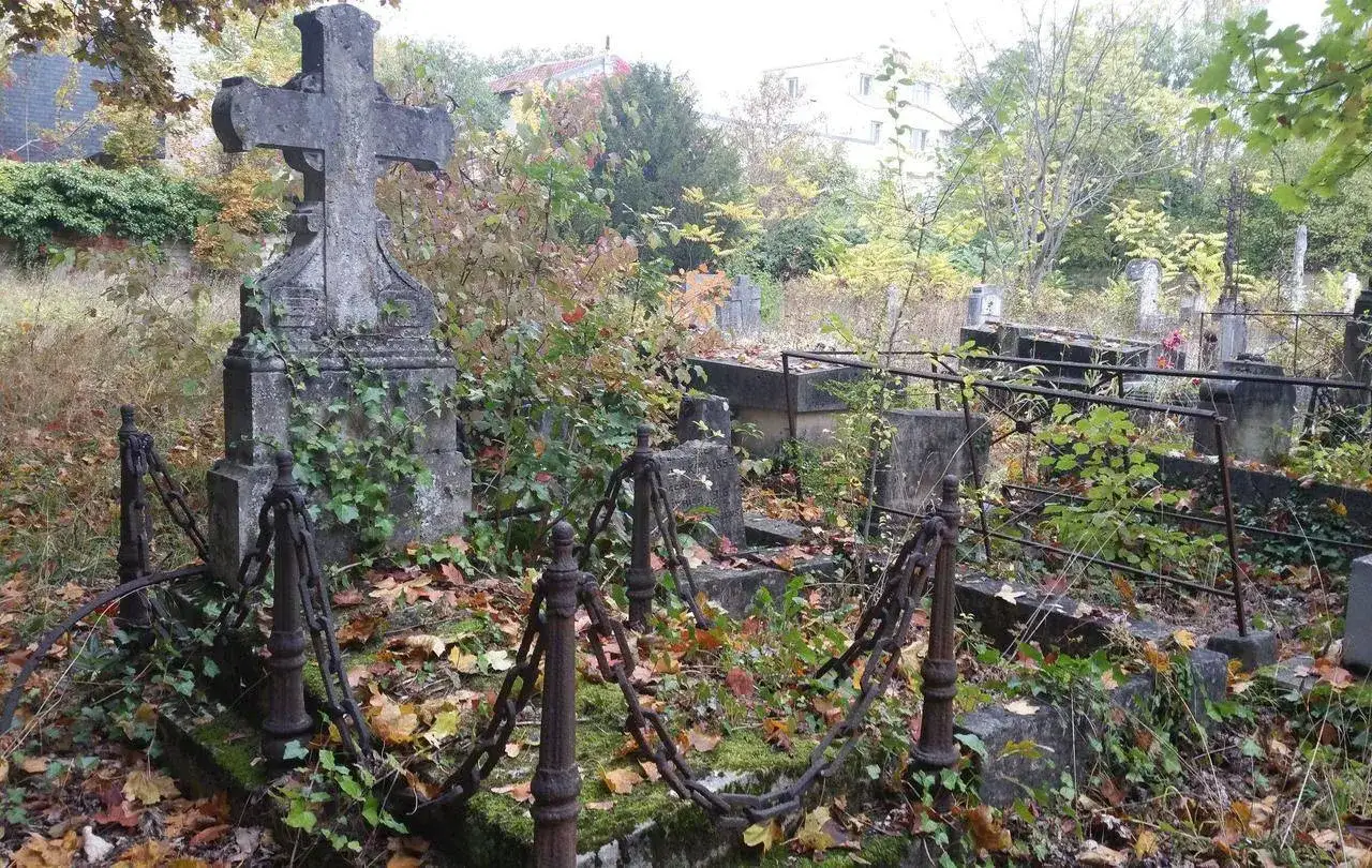 Un cimetière abandonné en France, envahi par la végétation. Une croix de pierre imposante se dresse au milieu des tombes oubliées, entourée de chaînes rouillées.