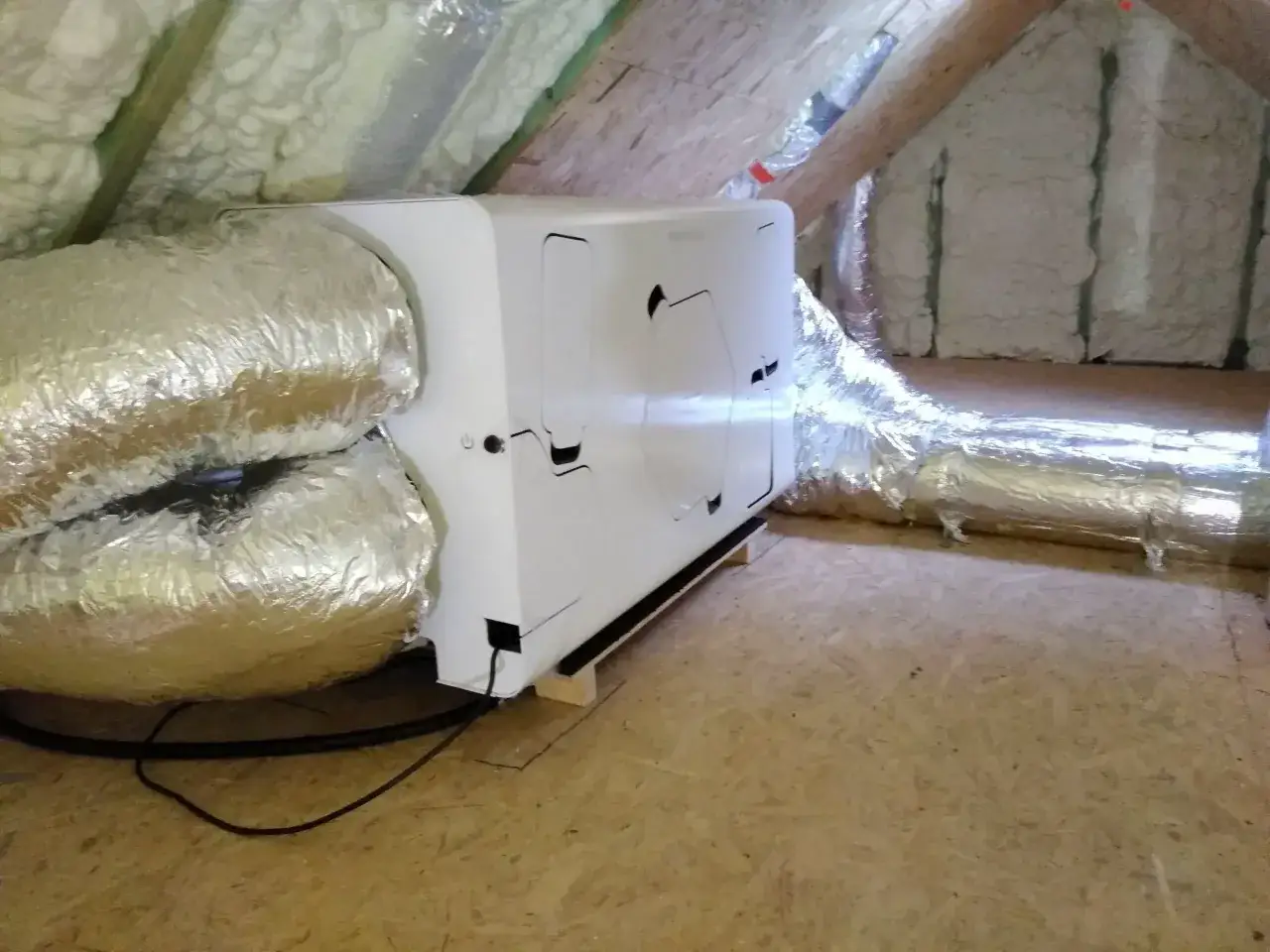Biały rekuperator z aluminiowymi kanałami wentylacyjnymi na nieogrzewanym poddaszu, otoczony izolacją i drewnianymi belkami.