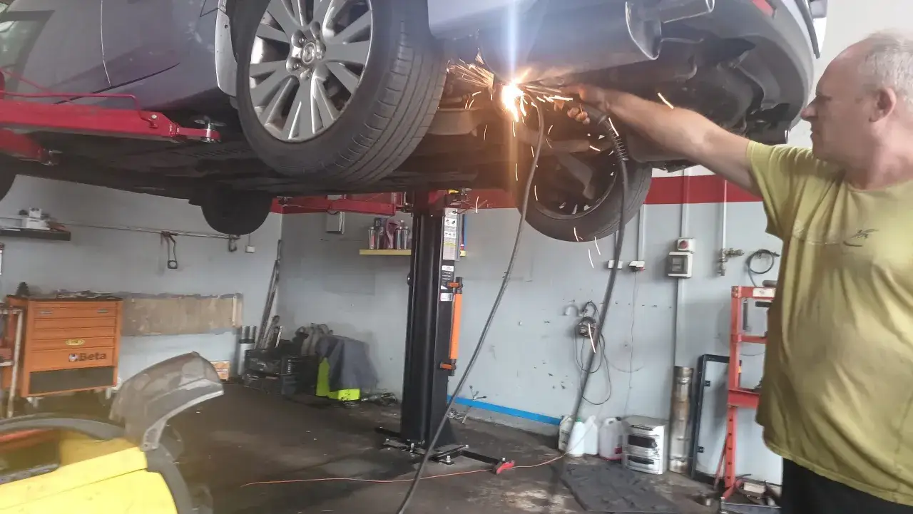 Mechanik wykonuje spawanie wydechu w podniesionym samochodzie. Iskry sypią się podczas pracy.