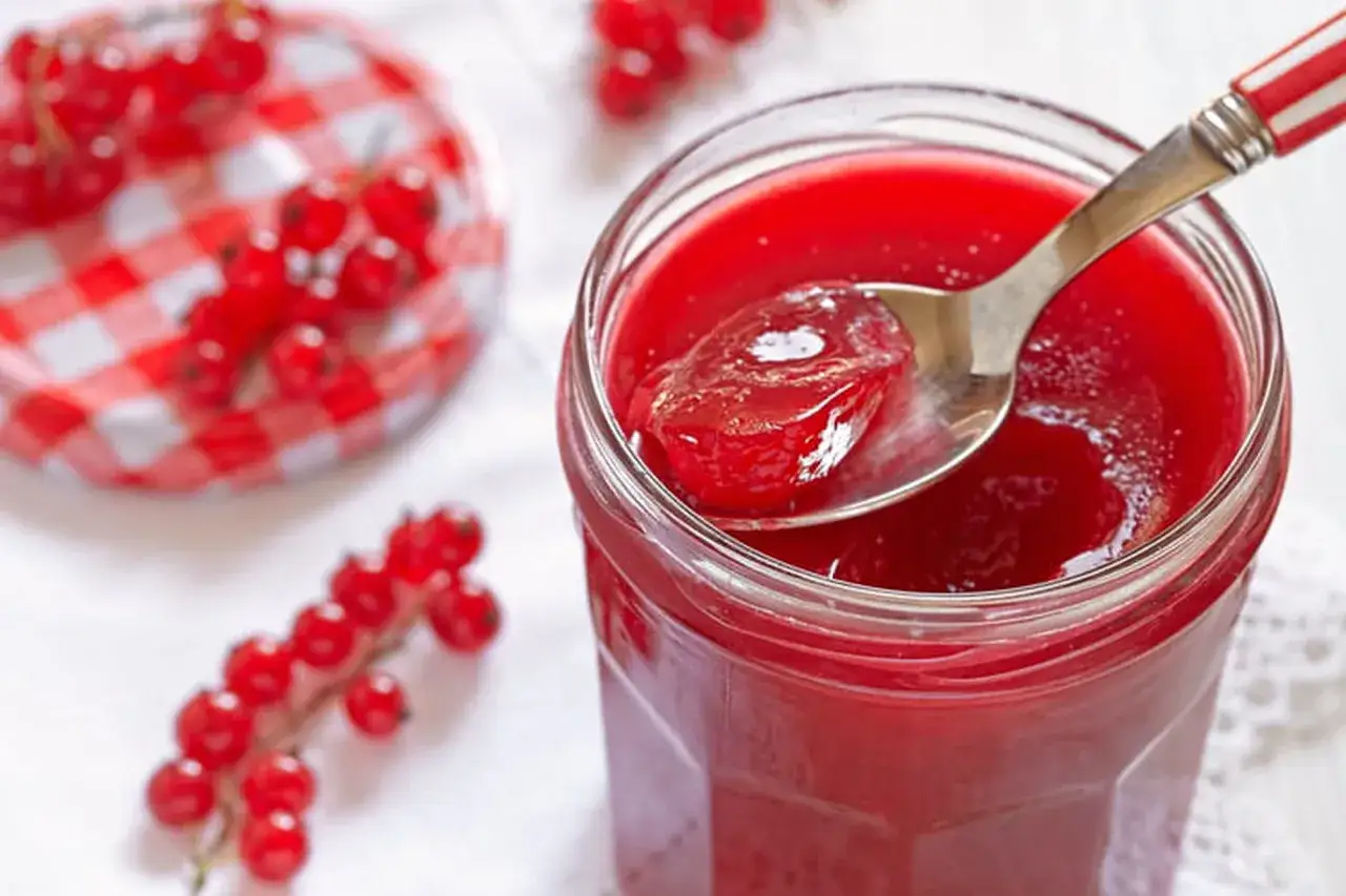 Une cuill&egrave;re de gelee de groseille, brillante et rouge, sort d'un pot. Des groseilles fra&icirc;ches sont dispos&eacute;es autour.