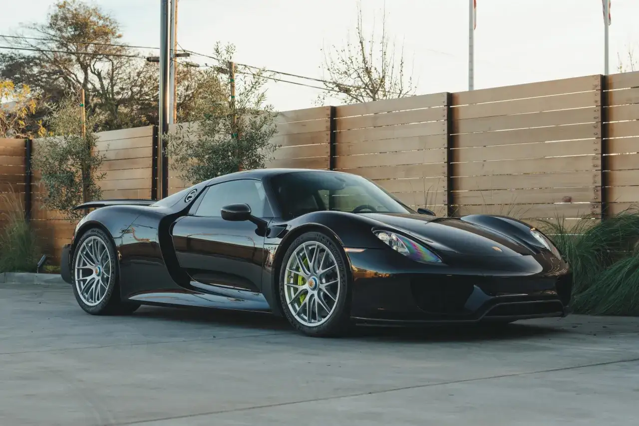 Czarny Porsche 918 Spyder lśni na tle drewnianego płotu i zieleni.