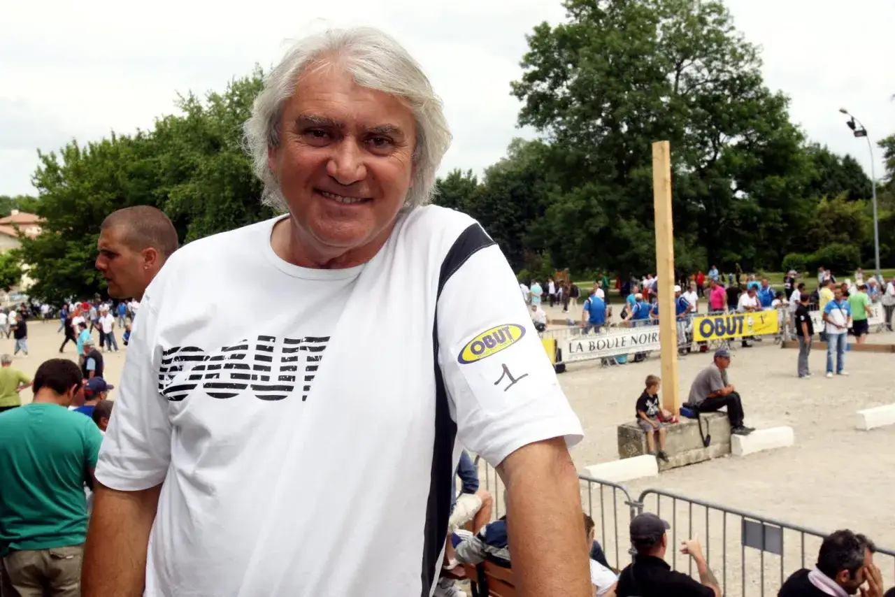 Un nom joueur de p&eacute;tanque fran&ccedil;ais souriant, portant un t-shirt blanc avec le logo OBUT. En arri&egrave;re-plan, un terrain de p&eacute;tanque anim&eacute;.