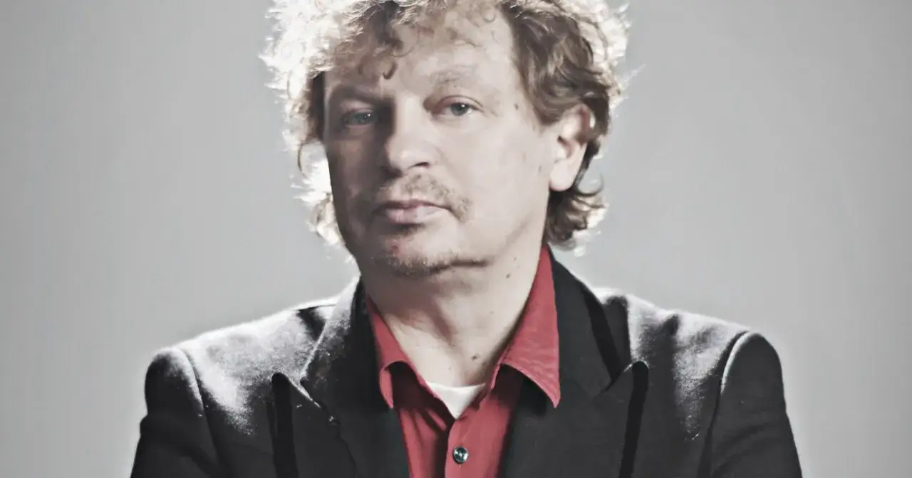 Marek Zieliński, aktor, w czarnej marynarce i czerwonej koszuli, z burzą kręconych włosów.