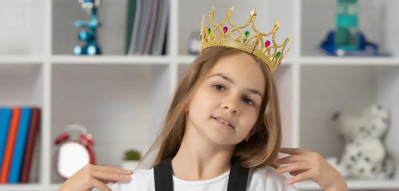 Mała egocentryczka w złotej koronie z kolorowymi kamieniami, pewna siebie, z uśmiechem na twarzy, na tle regału z książkami i zabawkami.