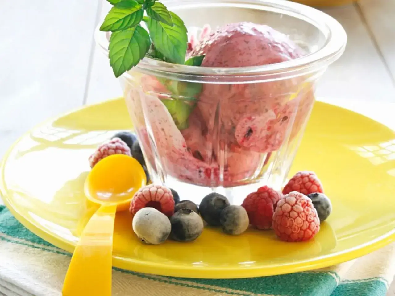 Fruchtiges Eis mit Sahne, garniert mit frischer Minze und Beeren. Ein gelber L&ouml;ffel liegt daneben.