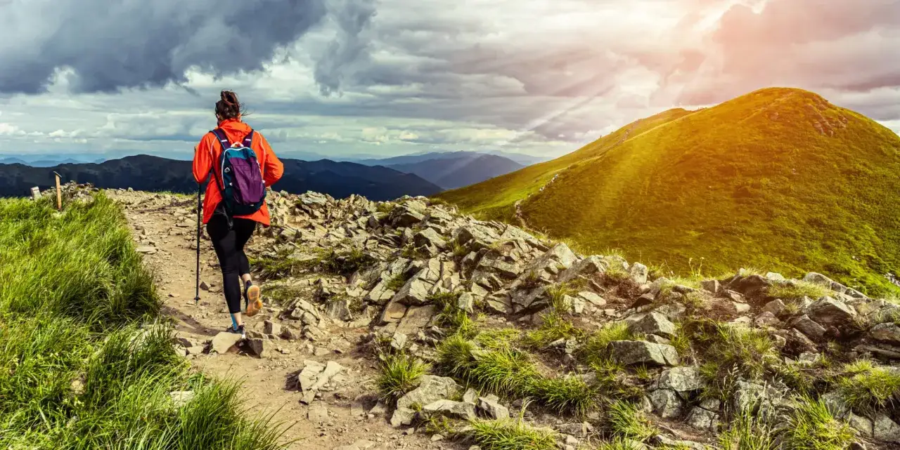 Kobieta w pomarańczowej kurtce wędruje po kamienistej ścieżce. W tle rozciągają się zielone wzgórza i góry, tworząc malownicze **trasy trekkingowe Polska**.
