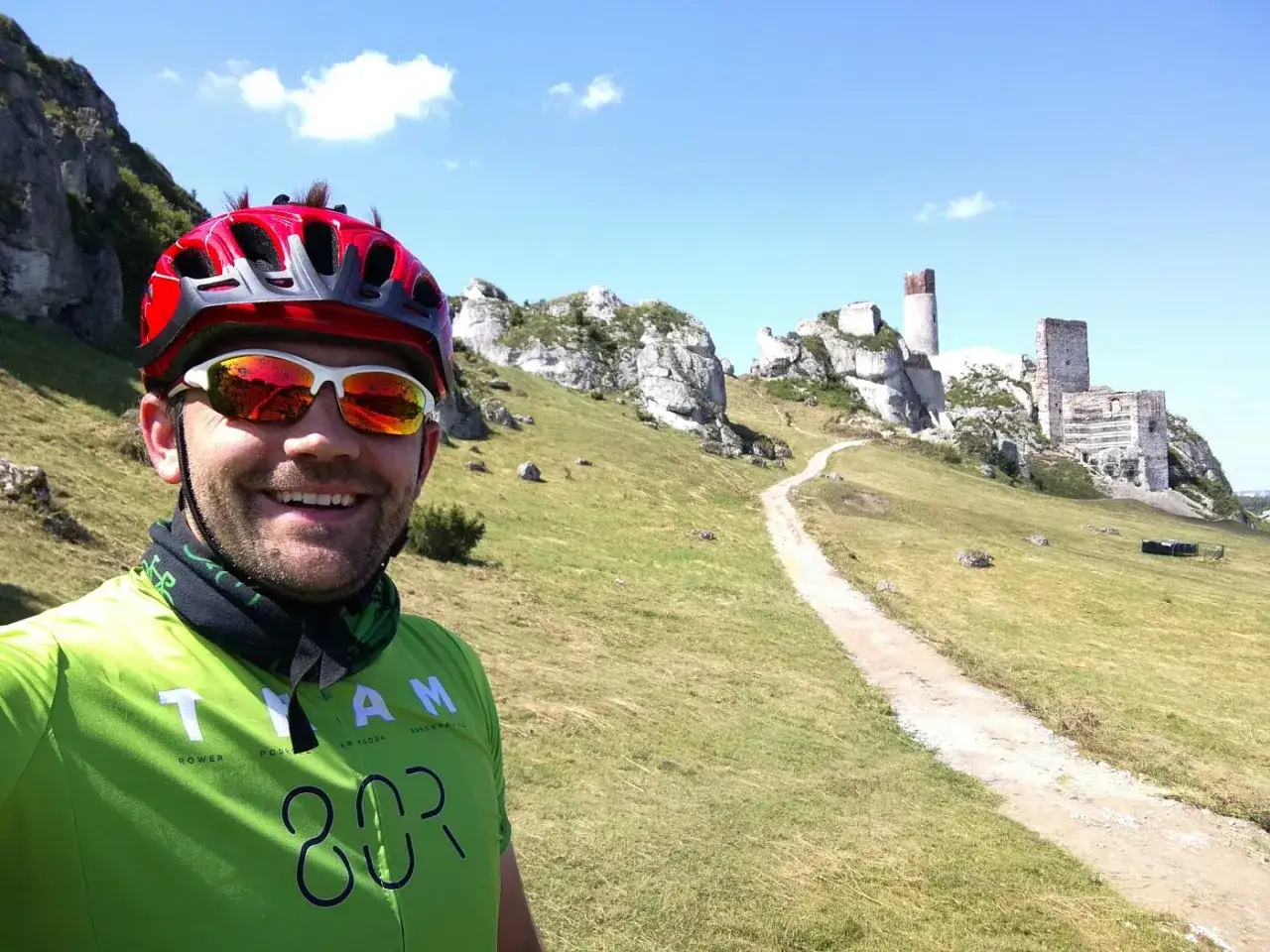 Uśmiechnięty rowerzysta w kasku i okularach na tle ruin zamku na Szlaku Orlich Gniazd.