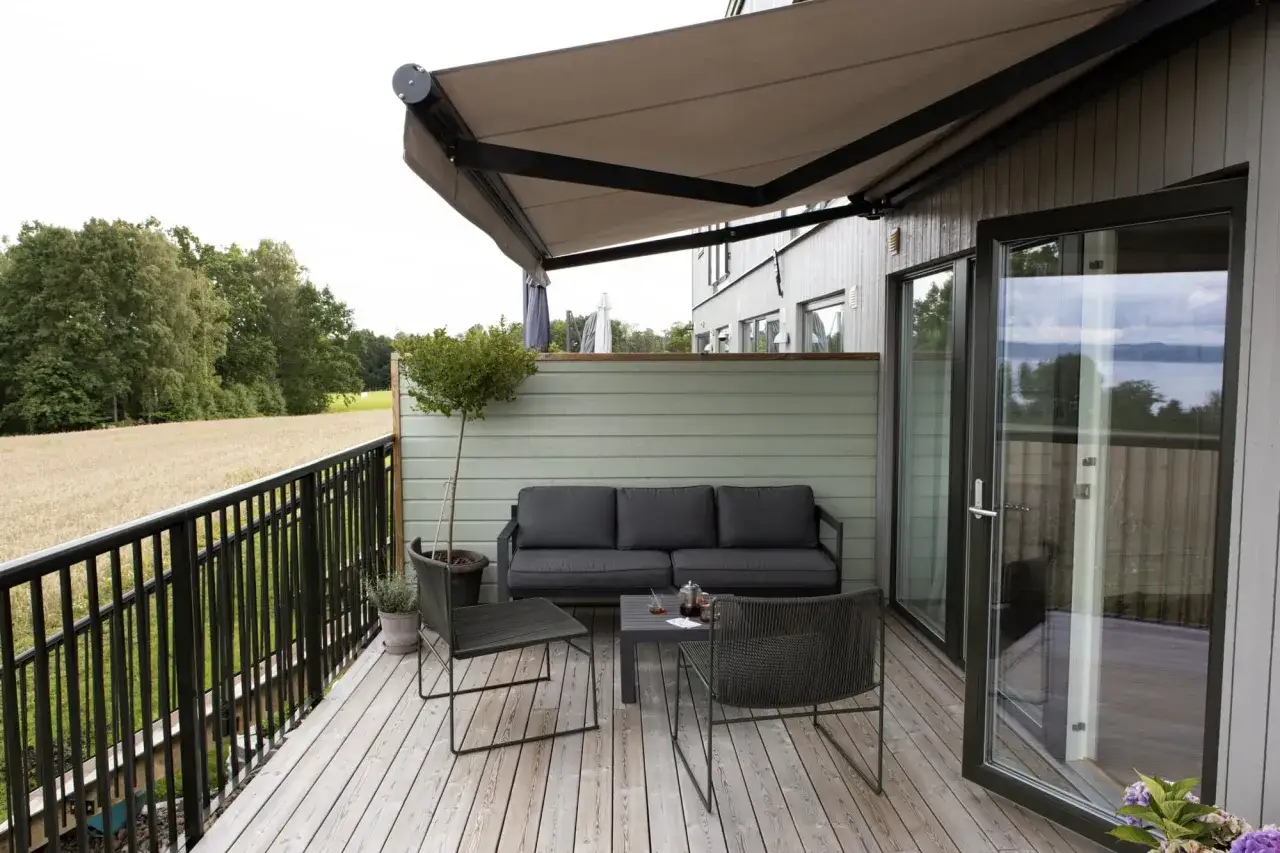 En uteplats med soffa, bord och stolar under en utdragen markis. Utsikt över fält och träd.