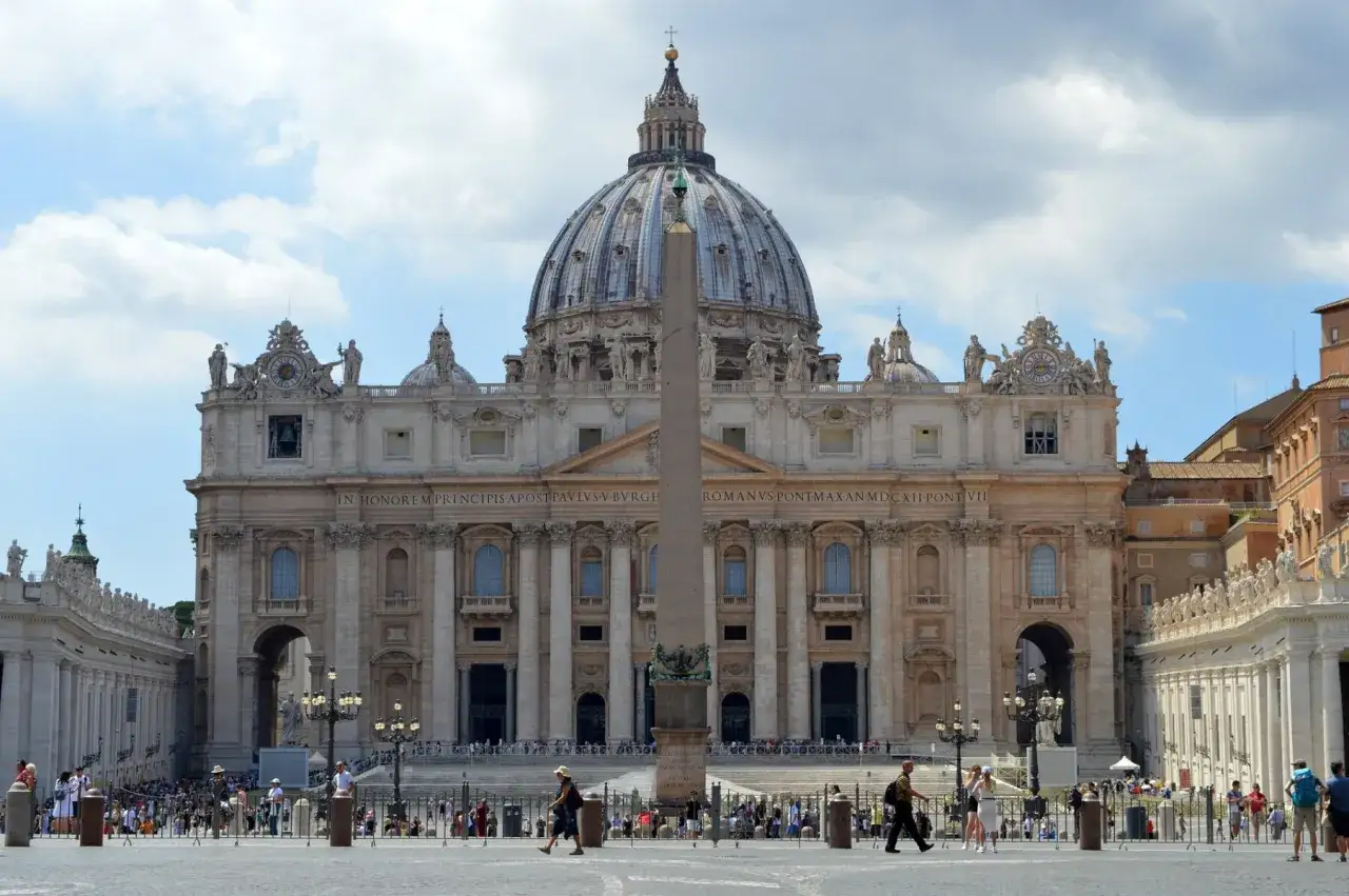 La maestosa facciata di San Pietro domina la piazza, con la sua cupola imponente e l'obelisco centrale. Turisti ammirano la grandezza.