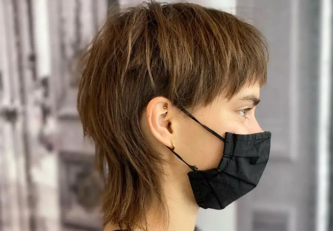 Frau mit kurzem Pixie Vokuhila-Haarschnitt, tr&auml;gt schwarze Maske und goldene Ohrringe.