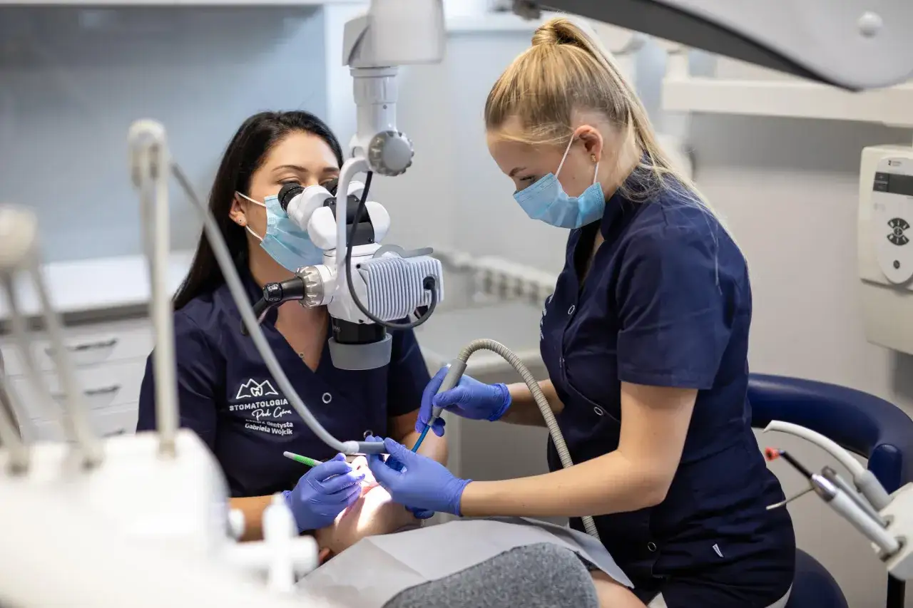 Dwie dentystki w maseczkach pracują przy pacjencie. Jedna używa mikroskopu, druga wierci. To profesjonalne leczenie, a nie domowa plomba do zęba.