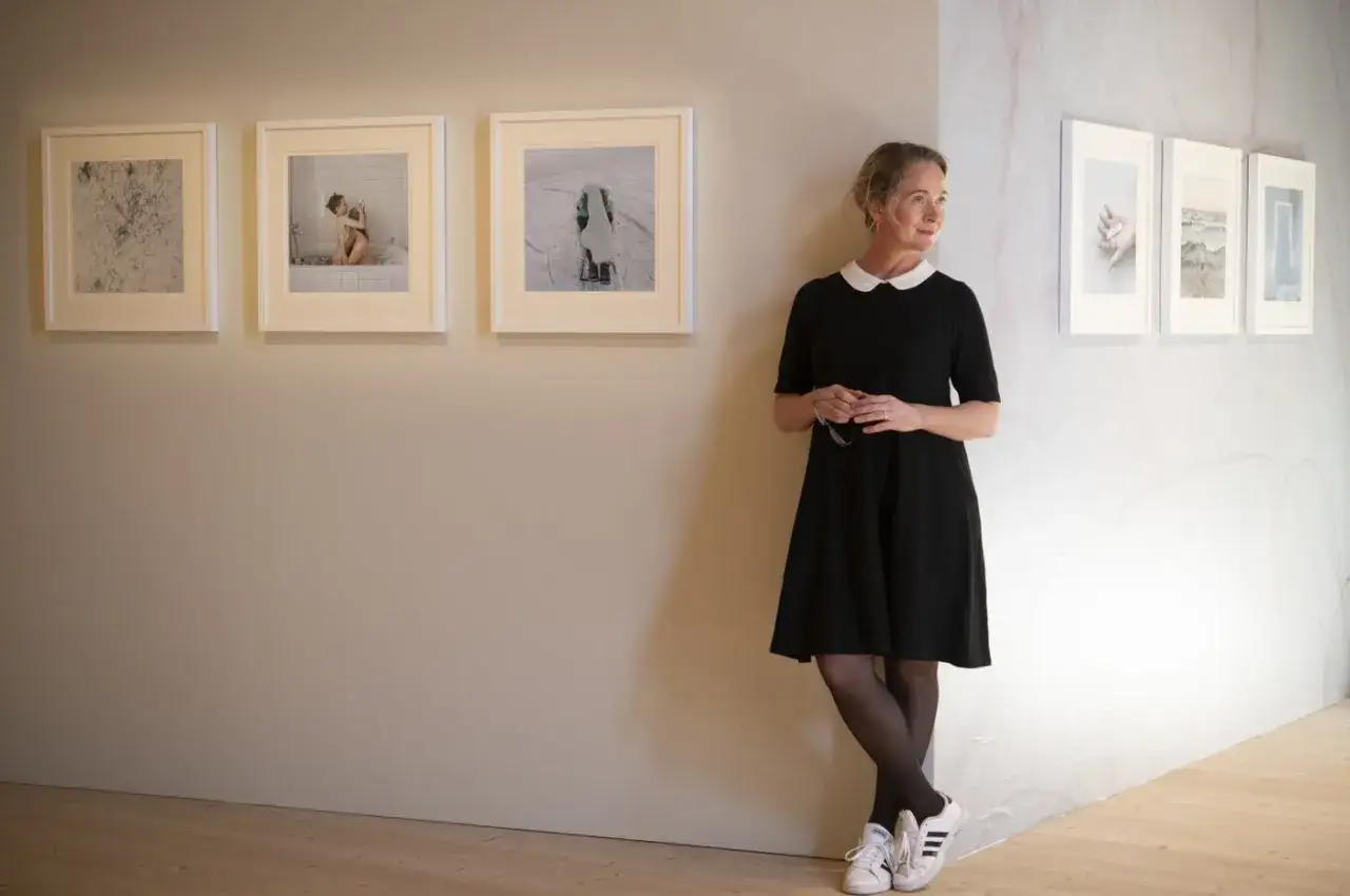 Anna Claren står vid en vägg med konstverk. Hon bär en svart klänning och tittar bort.