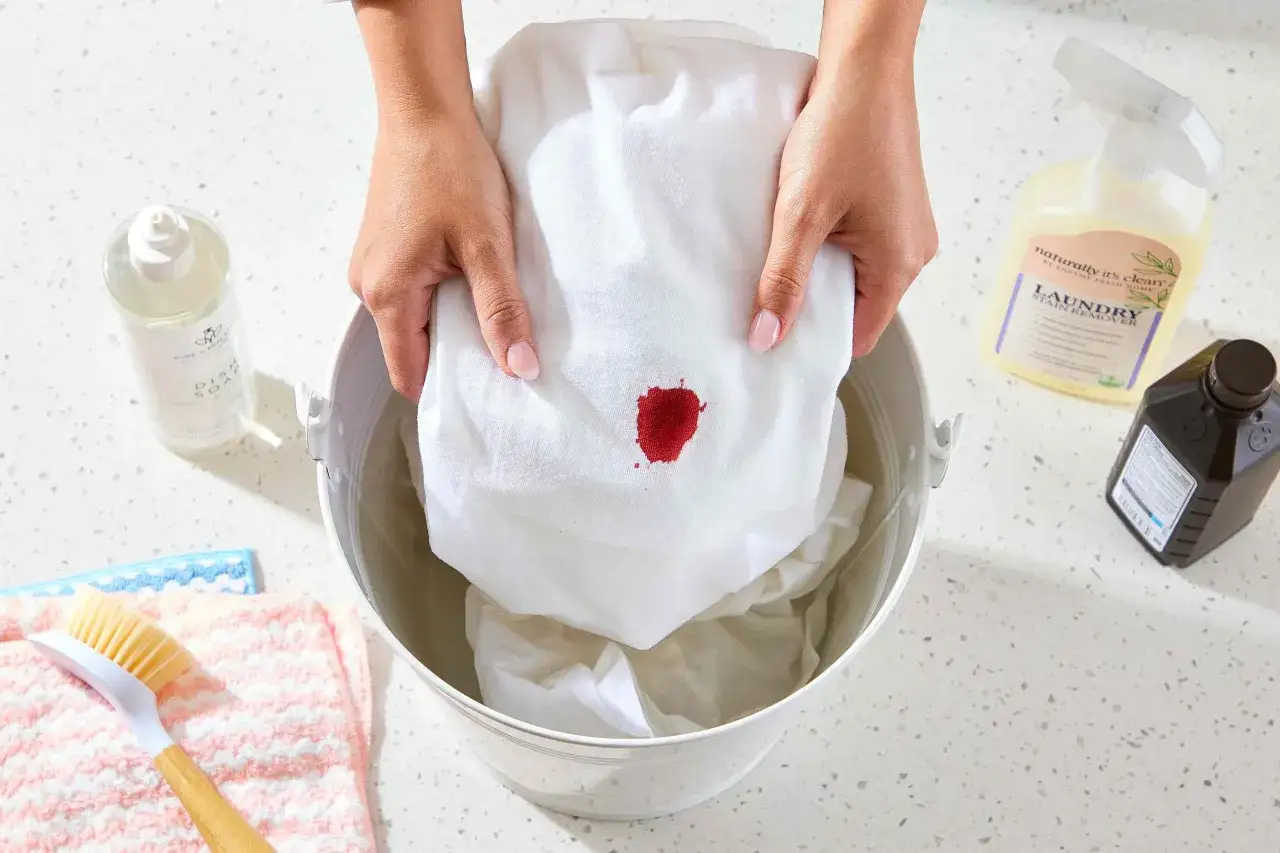 Manos sostienen tela blanca con una mancha roja, prepar&aacute;ndose para quitar manchas de sangre secas con productos de limpieza.