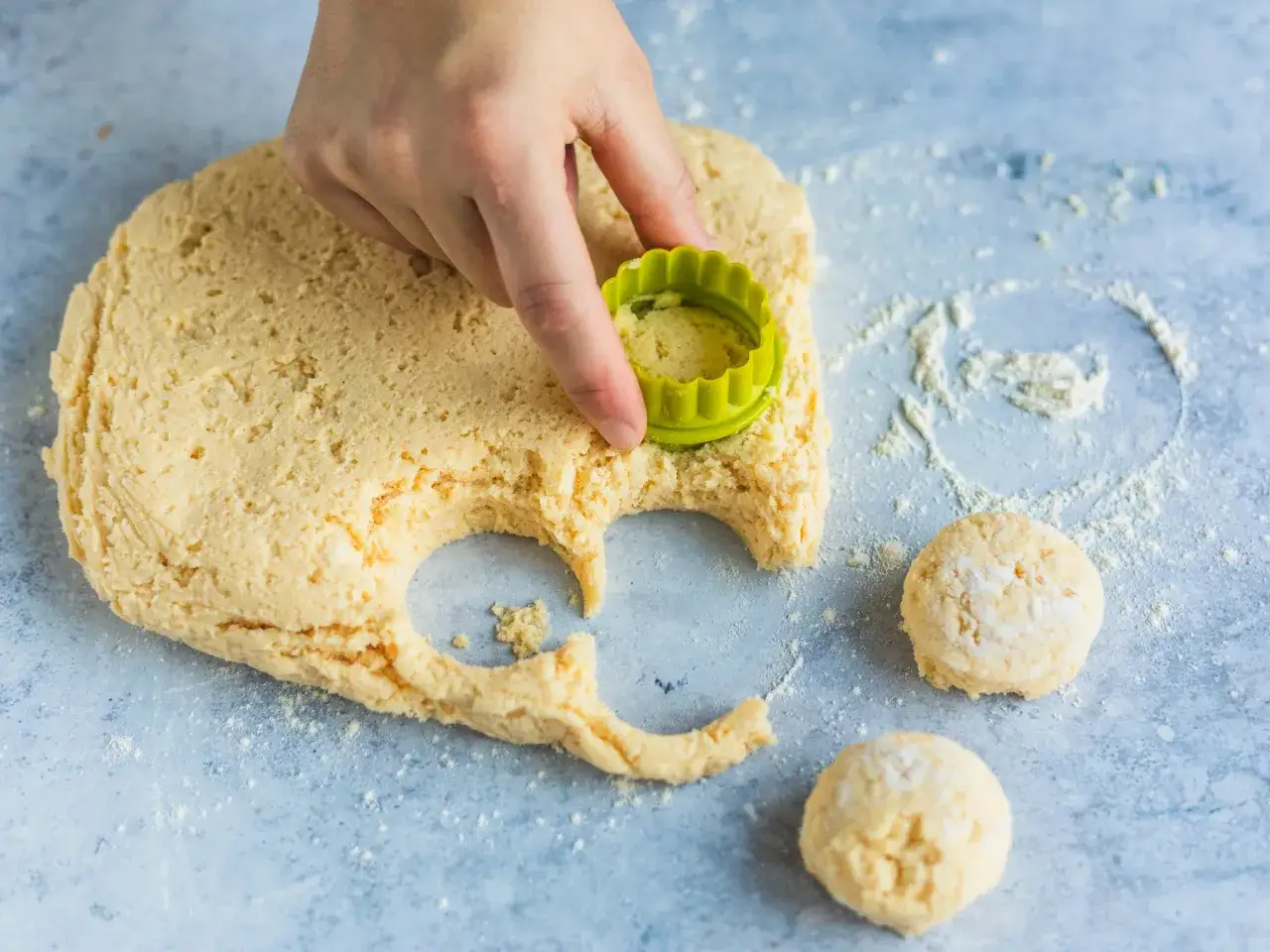 Hand formt mit grünem Ausstecher aus hellem Scones Teig runde Kekse. Zwei fertige Kekse liegen daneben.