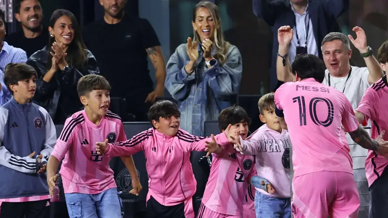 Messi w różowej koszulce z numerem 10, otoczony przez dzieci, które mogą być jego synem. W tle widać kibiców, w tym jego żonę.