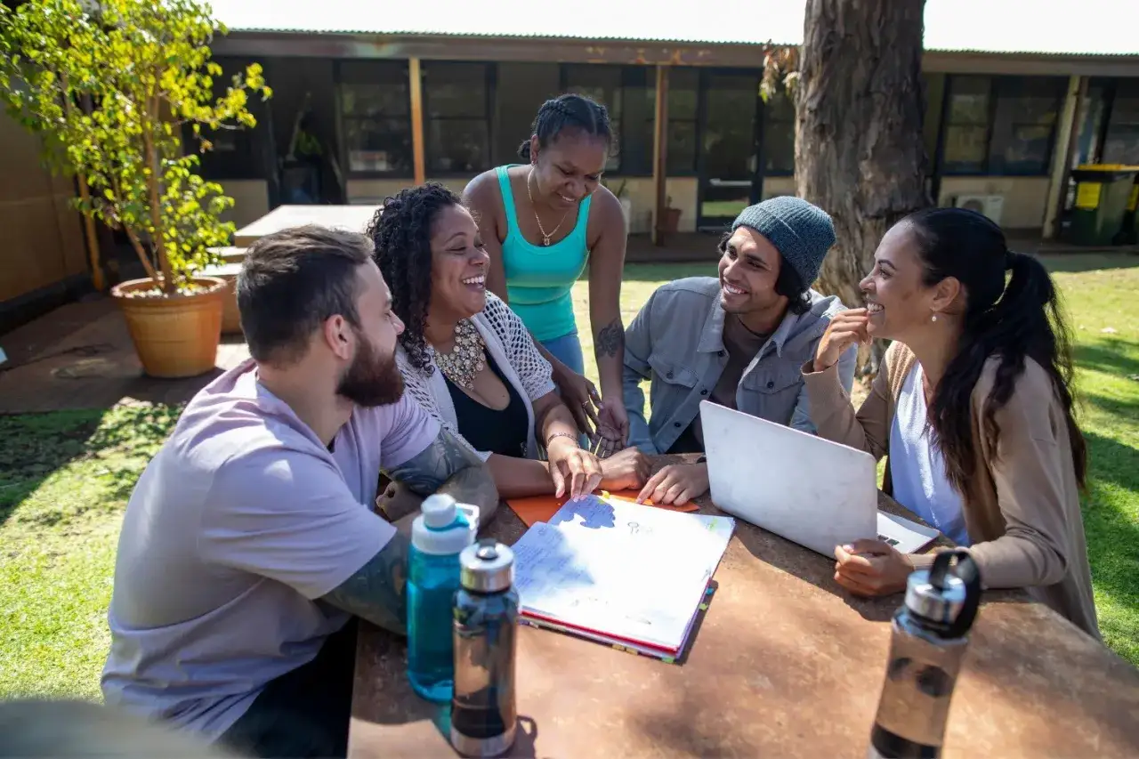 A diverse group of young adults collaborates outdoors. They share laughter and ideas, with a laptop and papers on the table, embodying a spirit where leaders speak last.