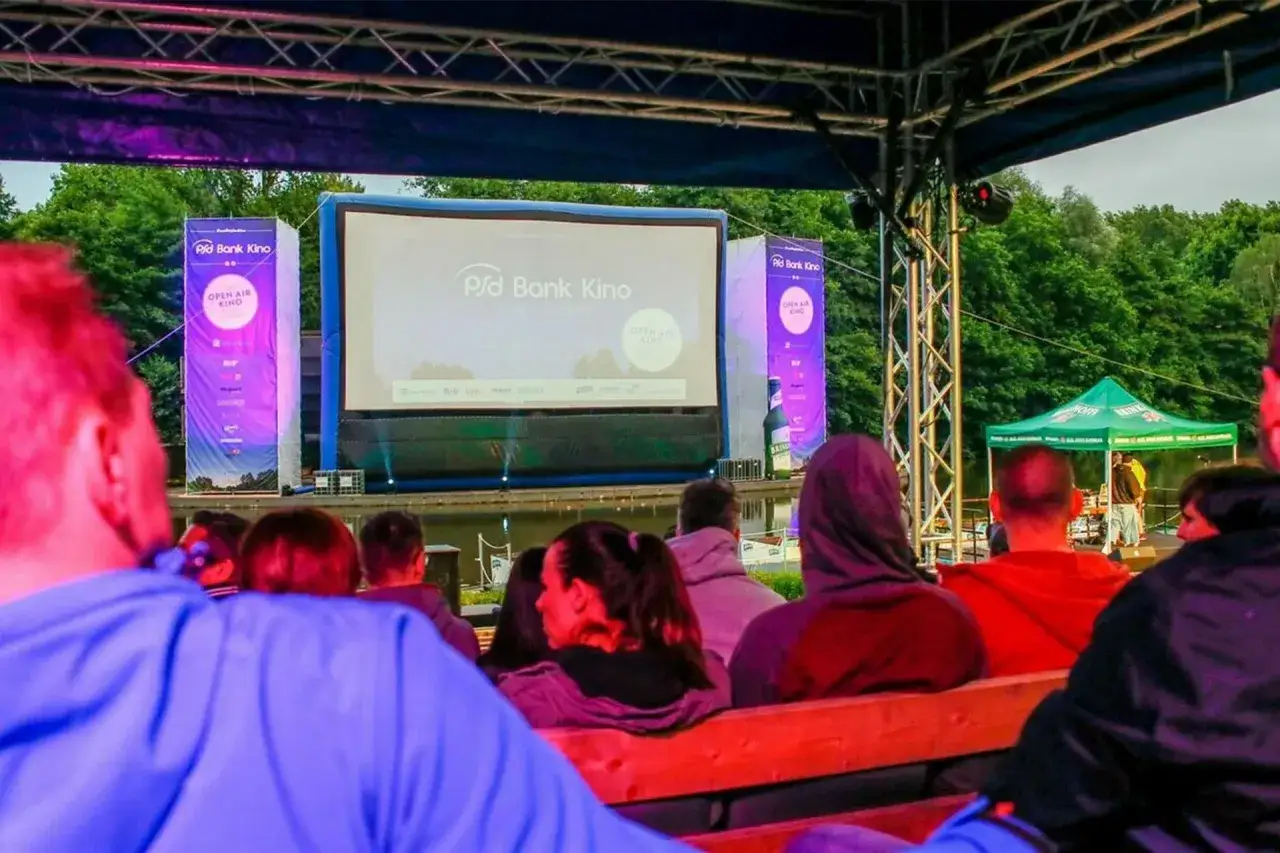 Menschen sitzen auf Holzb&auml;nken und genie&szlig;en einen Film im **freiluft kino**. Eine gro&szlig;e Leinwand mit dem Logo 