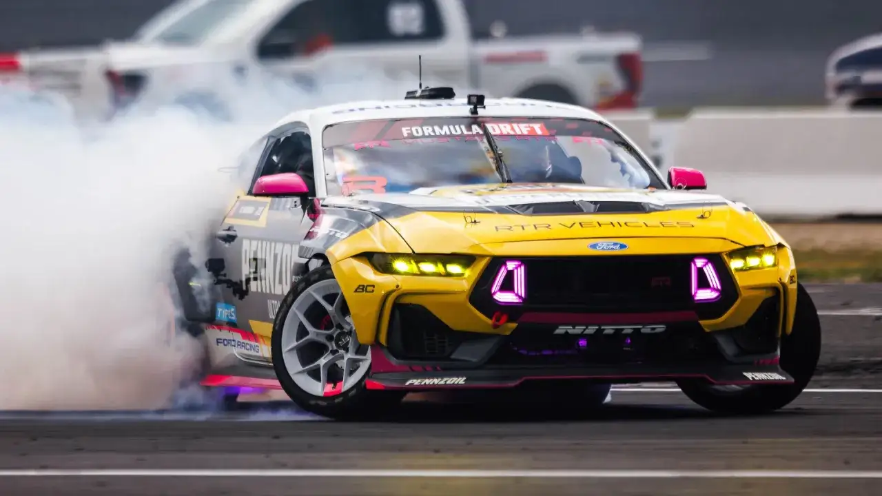 Żółty samochód do driftu Ford Mustang z różowymi akcentami i neonowymi światłami, wyrzucający kłęby dymu podczas zawodów Formula Drift.