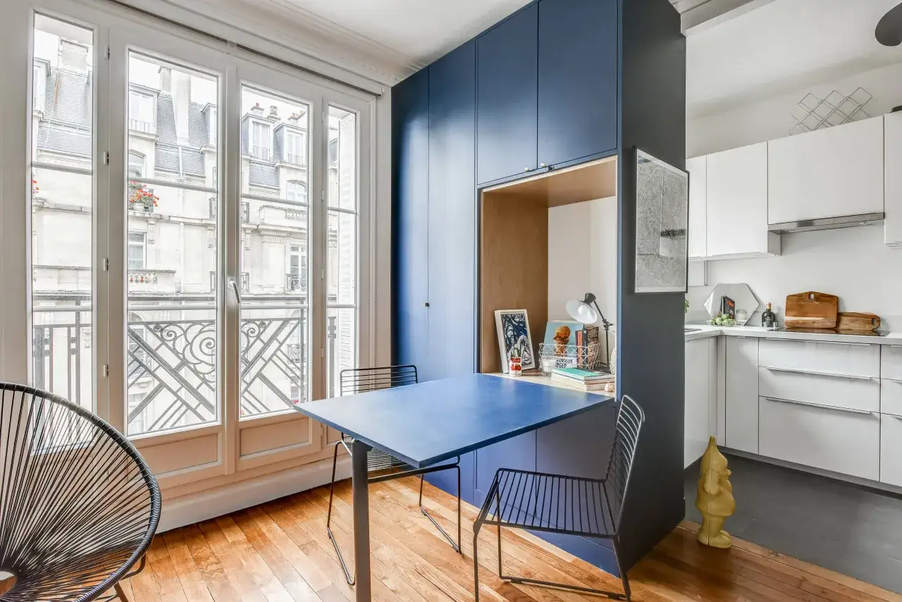 Aménagement petit studio : table bleue, chaises design, et rangements intégrés. Une cuisine blanche et une vue parisienne complètent l'ensemble.