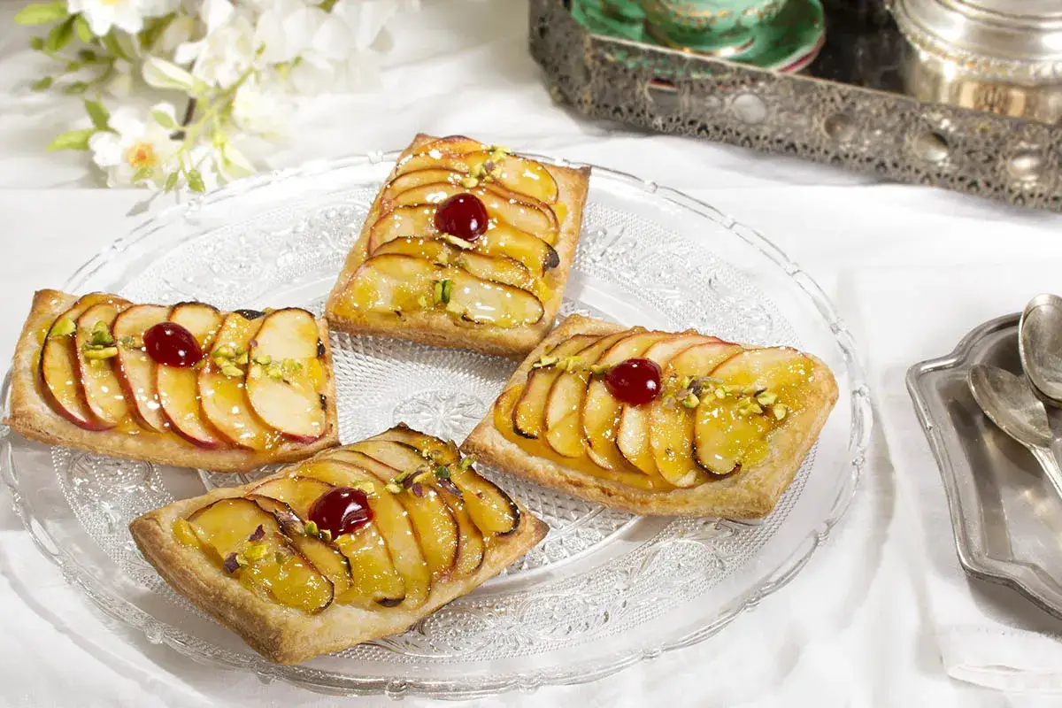 Cuatro tartaletas de manzana glaseadas, decoradas con pistachos y cerezas, servidas en una bandeja de cristal.