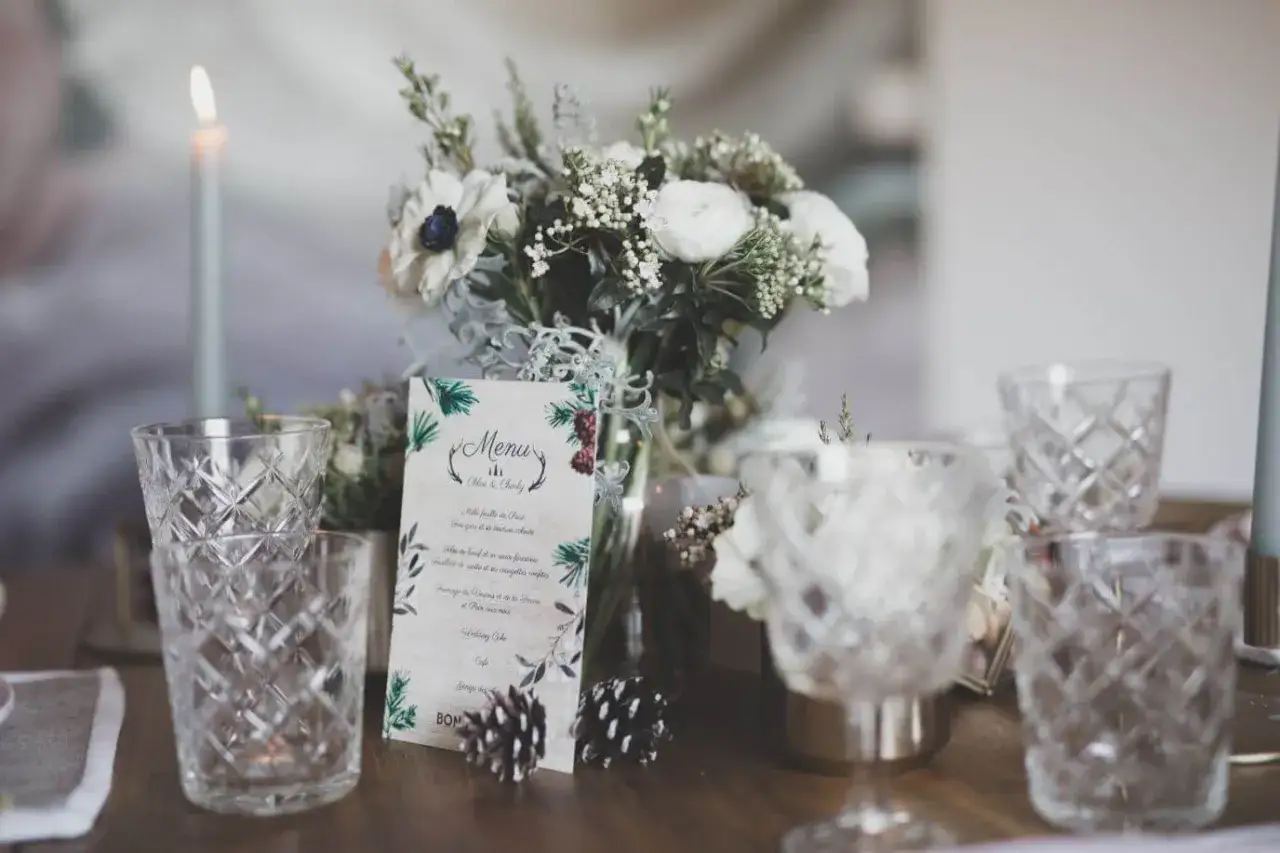 Un menu de mariage pour Chlo&eacute; & Charly, orn&eacute; de feuillages, est pos&eacute; sur une table dress&eacute;e avec des verres textur&eacute;s et un bouquet de fleurs blanches.