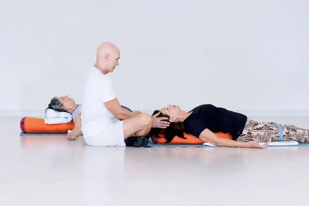 Un instructor de yoga asiste a dos mujeres en posturas restaurativas, demostrando los beneficios del yoga para la relajación y el bienestar.