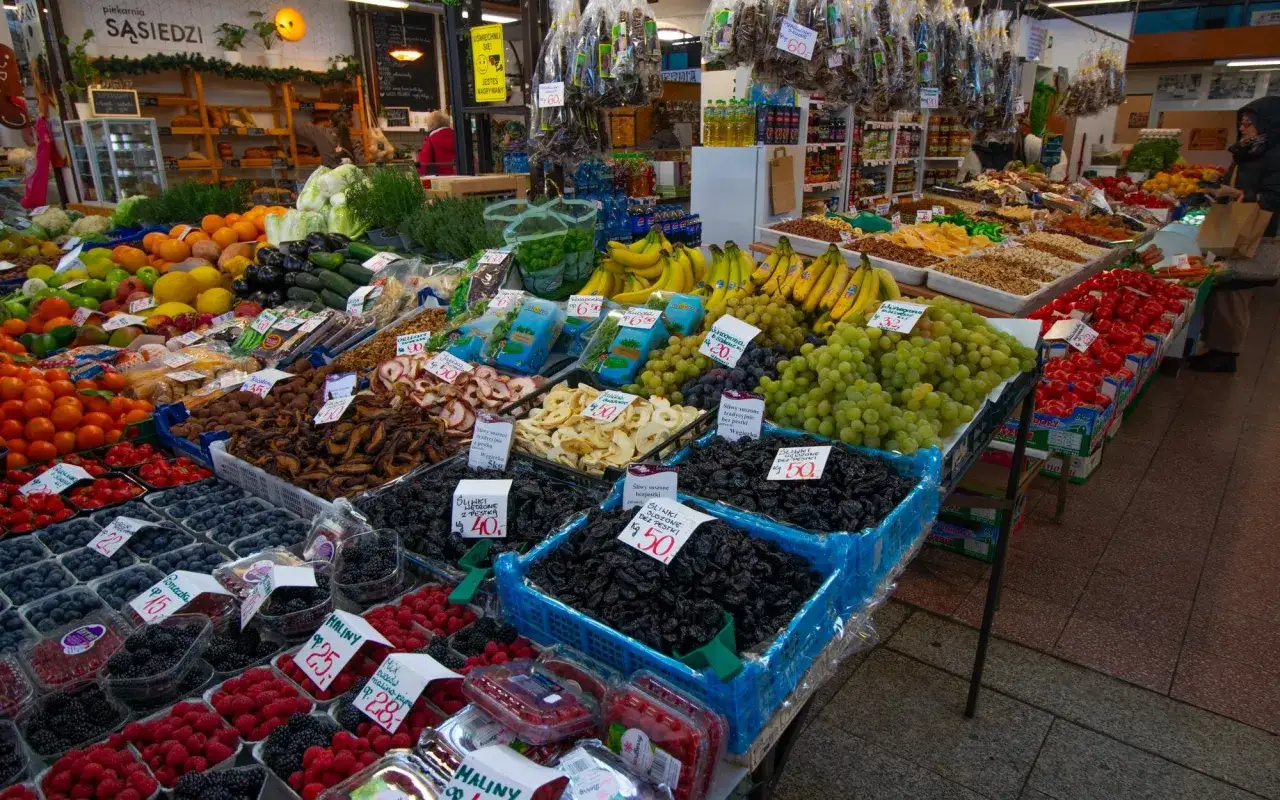 Targowisko pełne owoców i warzyw, kiedy sezon na truskawki. Widać maliny, borówki, winogrona, banany, pomarańcze i suszone owoce.