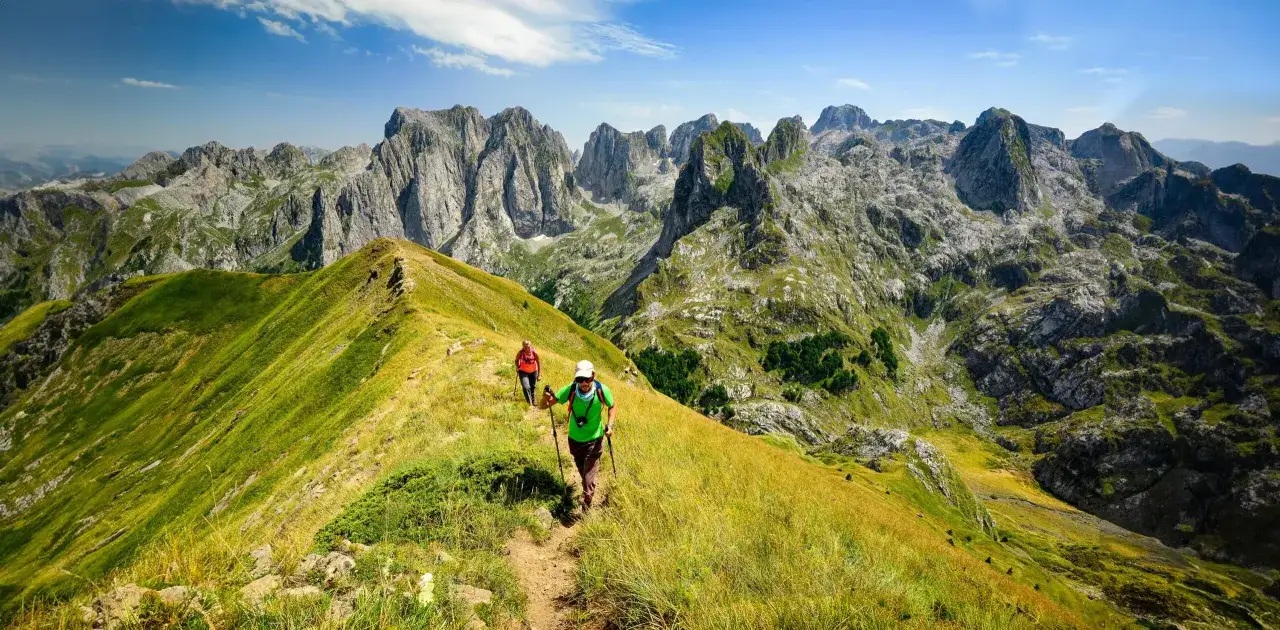 Deux randonneurs profitent d'un trek en Albanie, traversant une cr&ecirc;te herbeuse avec des montagnes majestueuses en arri&egrave;re-plan.