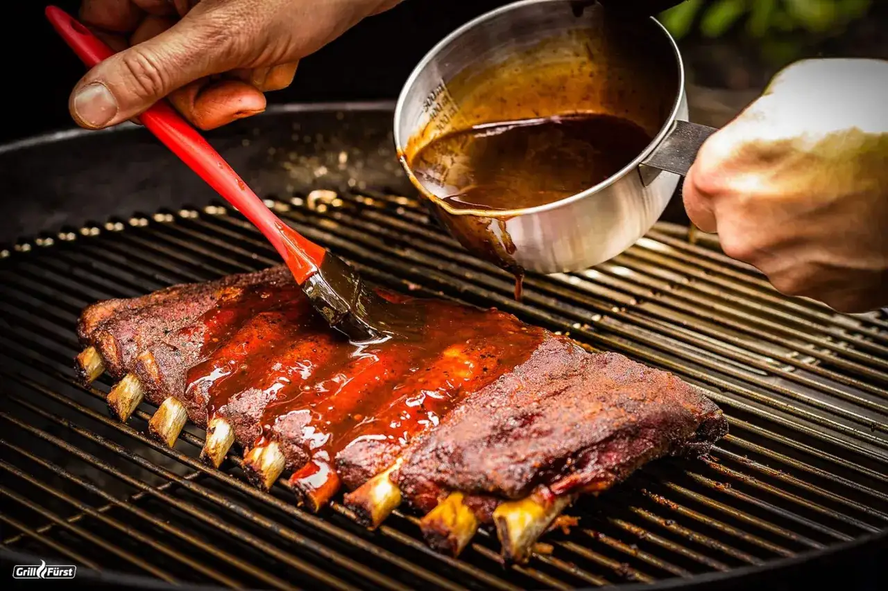 Rippchen einlegen zum Grillen: Eine Hand bestreicht saftige Rippchen mit einer gl&auml;nzenden BBQ-Sauce, w&auml;hrend eine andere Hand die Sauce aus einem Messbecher nachgie&szlig;t.