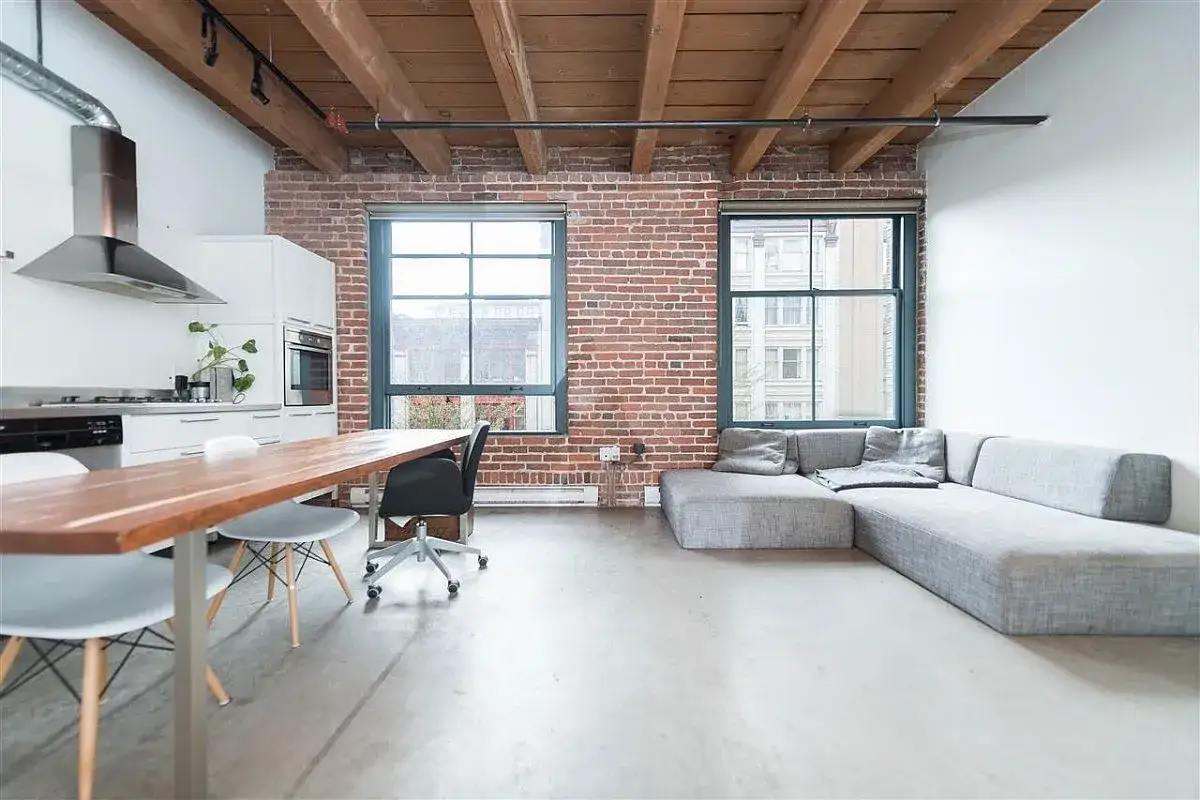 Nowoczesne loft mieszkanie z ceglaną ścianą, drewnianym sufitem, kuchnią, stołem i szarą kanapą.