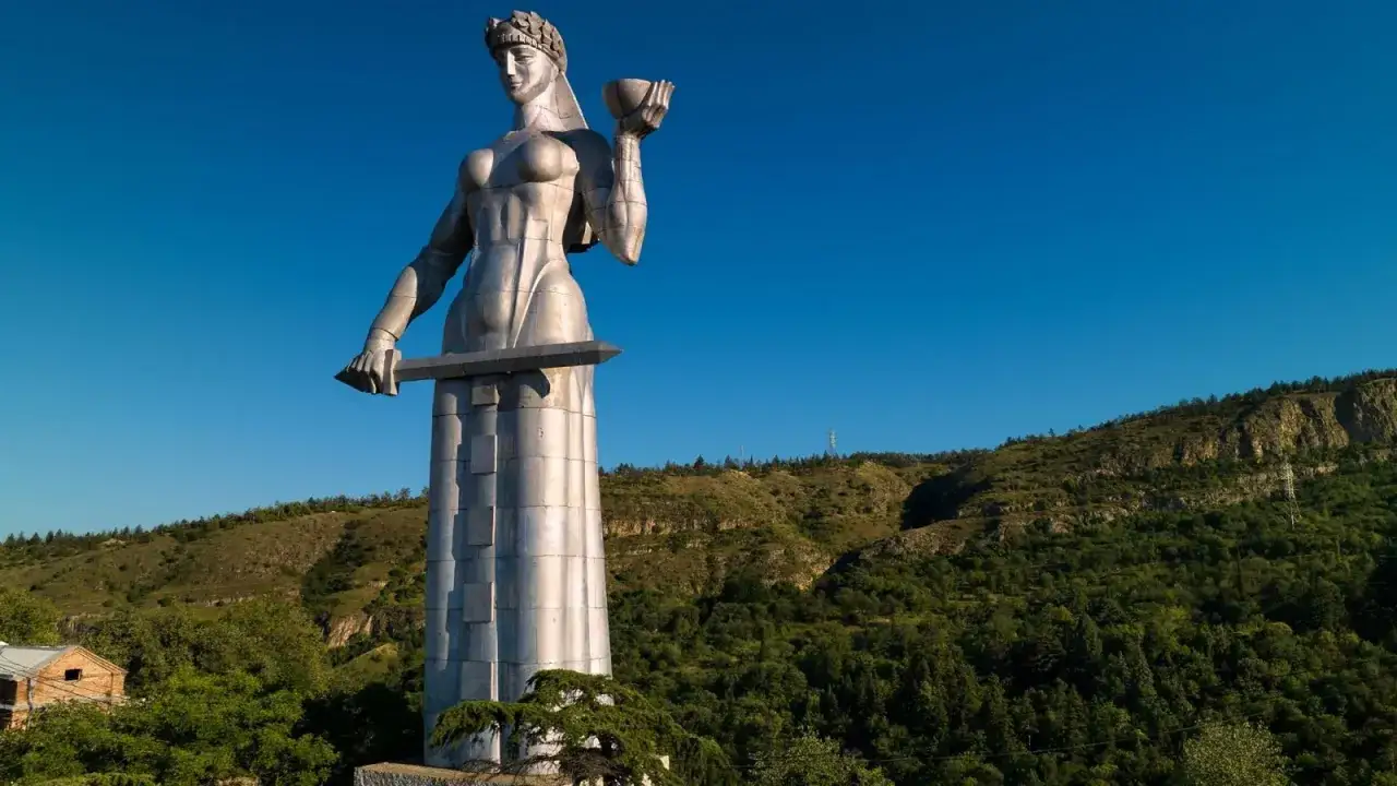 Matka Gruzja, symbol gościnności i siły, wznosi się nad wzg&oacute;rzami. W jednej dłoni trzyma puchar wina, w drugiej miecz.
