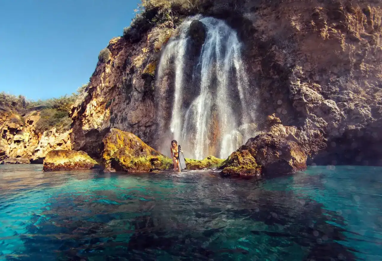 Una persona se encuentra en la cala del barranco de Maro, disfrutando de una cascada que cae sobre rocas y el mar turquesa.